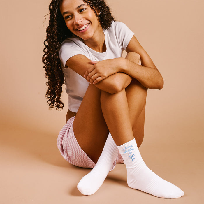 A young woman with curly dark hair sits cross-legged, wearing a white t-shirt, pink striped shorts, and The Darling Effect's Alpha Delta Pi Embroidered Crew Socks—white socks with blue bow and text—showing true sorority spirit.