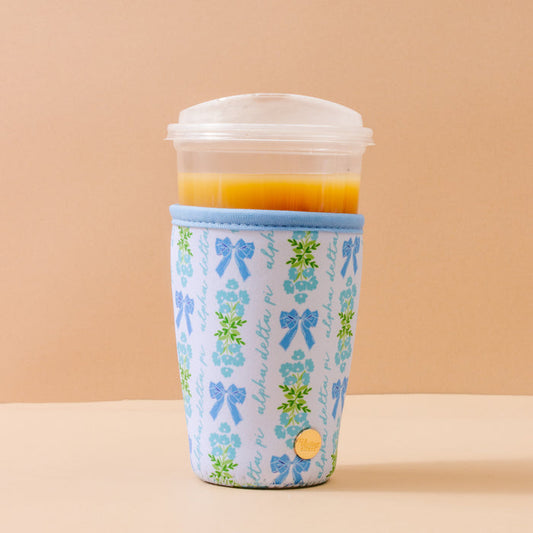 An Alpha Delta Pi Large Sip Savvy Drink Sleeve by The Darling Effect, featuring blue bows and green leaves, wraps a clear plastic cup of orange juice with a lid. The cup sits on a beige surface against a beige background.