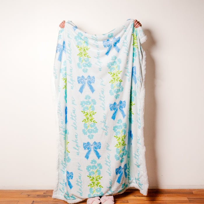 The Darling Effect's Alpha Delta Pi Plush Sherpa Blanket features a white base with light blue flowers, green leaves, blue bows, and "Alpha Delta Pi" in script—ideal as a cozy sorority blanket or stylish college dorm throw.