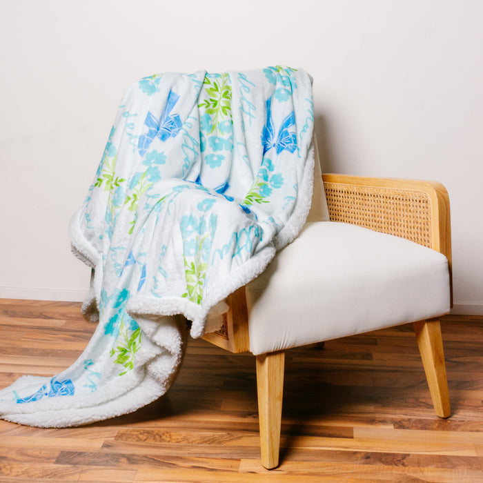 A white armchair with light wood and cane paneling is draped with The Darling Effect's Alpha Delta Pi Plush Sherpa Blanket, featuring blue and green floral and bird patterns, on a wood floor against a plain white wall.