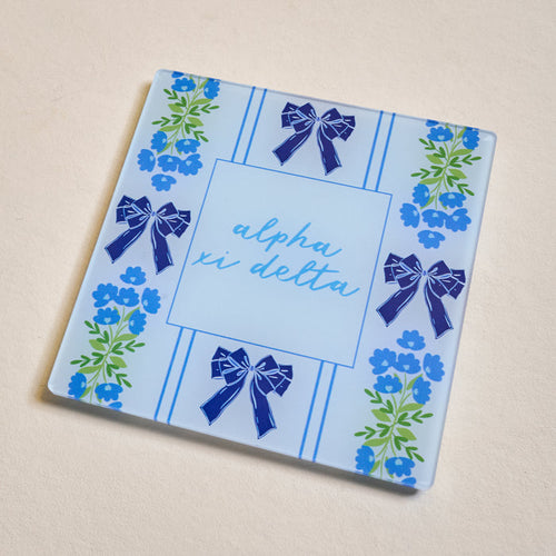The Darling Effect's Alpha Xi Delta Acrylic Coaster features a square blue design with dark blue bows, green-and-blue floral edge patterns, and "alpha xi delta" in light blue script—an ideal sorority gift.