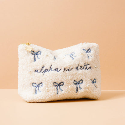 The Darling Effect’s Alpha Xi Delta Bows & Pearls Teddy Pouch features cream sherpa, blue bow and script embroidery, pearl accents, a gold zipper pull with charm—a fuzzy, stylish sorority pouch perfect for gifting.