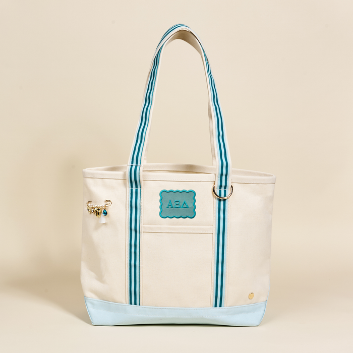 The Darling Effect's Alpha Xi Delta Canvas Tote Bag features beige canvas, blue striped handles, a blue Greek-letter patch, gold and blue charm, and light blue base—making it the perfect stylish sorority gift.
