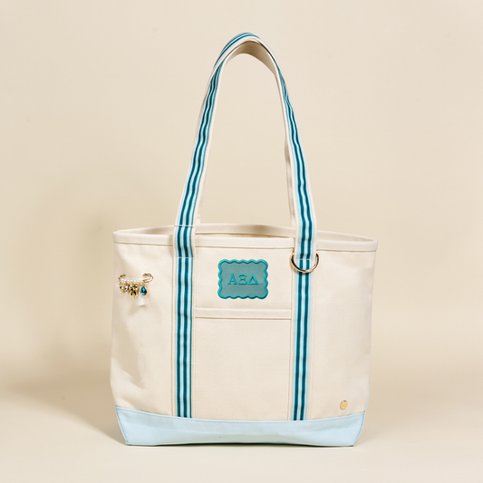 The Darling Effect's Alpha Xi Delta Canvas Tote Bag features beige canvas, blue striped handles, a blue Greek-letter patch, gold and blue charm, and light blue base—making it the perfect stylish sorority gift.