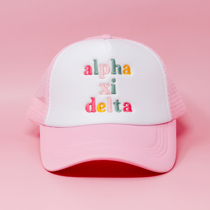 The Darling Effect’s Alpha Xi Delta Pink Trucker Hat features colorful embroidered letters on the front and is shown against a pink background.