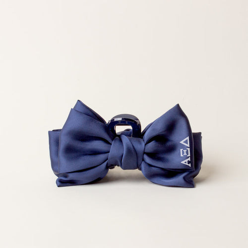 The Darling Effect's Alpha Xi Delta Satin Bow Claw Clip features a navy satin bow, silver claw clip, and white ΑΕΔ letters—perfect for updos and shown on a plain light background.