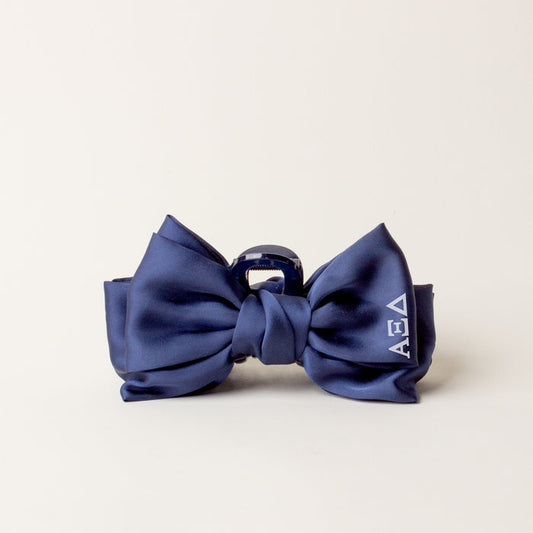 The Darling Effect's Alpha Xi Delta Satin Bow Claw Clip features a navy satin bow, silver claw clip, and white ΑΕΔ letters—perfect for updos and shown on a plain light background.