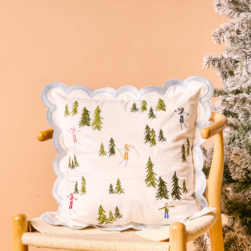 The Darling Effect's Aprés Ski Scalloped Square Throw Pillow features a white cover with a blue scalloped border, embroidered evergreen trees and skiers—perfect for lodge decor beside a frosted Christmas tree.