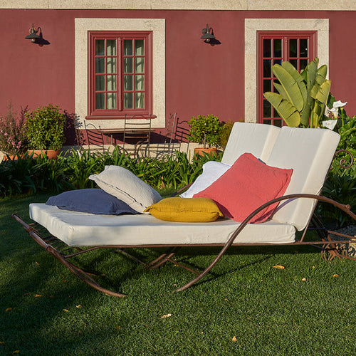 White lounge chair with Areia saffron white and terracotta decorative pillowcases on a grassy area in front of a red building.