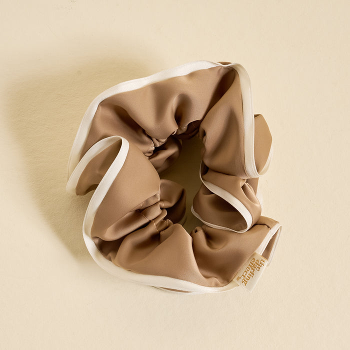 The Darling Effect's Beige Ruffle Up Scrunchie, an oversized tan satin hair accessory with white trim and a small fabric tag, is displayed in a circular shape on a light beige background for a stylish and gentle hold.