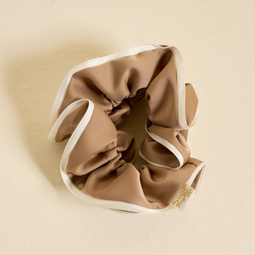 The Darling Effect's Beige Ruffle Up Scrunchie, an oversized tan satin hair accessory with white trim and a small fabric tag, is displayed in a circular shape on a light beige background for a stylish and gentle hold.