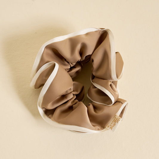 The Darling Effect's Beige Ruffle Up Scrunchie, an oversized tan satin hair accessory with white trim and a small fabric tag, is displayed in a circular shape on a light beige background for a stylish and gentle hold.