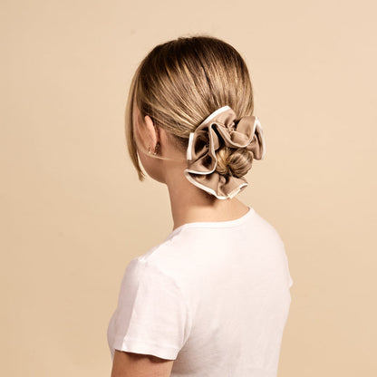 A woman with blonde hair in a low bun, styled with The Darling Effect's Beige Ruffle Up Scrunchie for a gentle hold, wears a white t-shirt and faces away from the camera against a plain beige background.