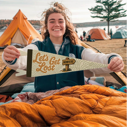 Camper with Benoit Let’s Get Lost 8x28.5” pennant at scenic lakeside campsite, multiple tents in outdoor adventure setting.