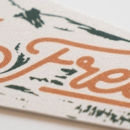 Benoit’s “Roam Wild & Free” felt pennant, 8x28.5”, features bold orange script and green vintage details, close-up view.