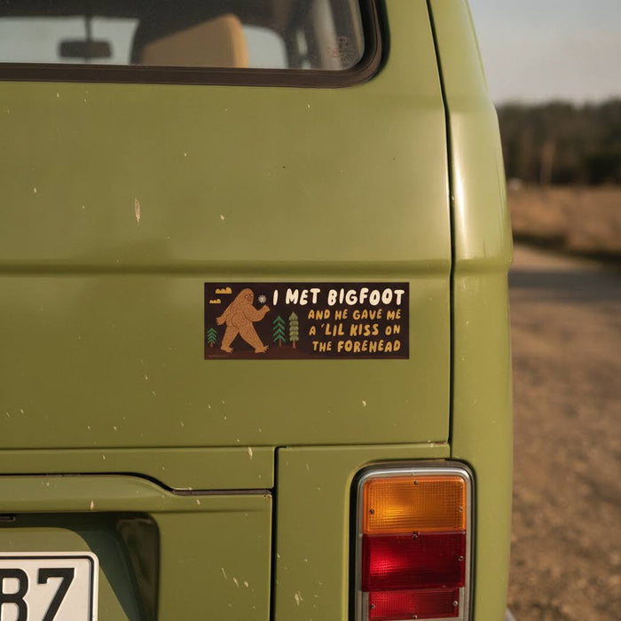 Green van with Betiobca “I Met Bigfoot and He Gave Me A Little Kiss” bumper sticker adds funny cryptid humor scene.