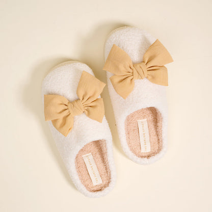 The Darling Effect's Betty Bow Tan Fuzzy Slippers, featuring plush white material, beige bows, and pink insoles, are displayed on a cream background.
