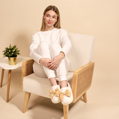 A young woman in white loungewear and The Darling Effect's Betty Bow Tan Fuzzy Slippers sits in a cream chair beside a small table with a potted plant.