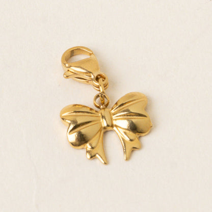 The Bitty Bow Jewelry Charm Gold by the-darling-effect features a shiny gold bow shape with a lobster clasp, ideal for necklaces or bracelets, displayed on an off-white background.