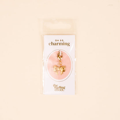 The Bitty Bow Jewelry Charm Gold by the-darling-effect comes attached to a card with “OH SO charming” and “the darling effect.”, all packaged in a clear plastic bag against a beige background—perfect for necklaces or bracelets.