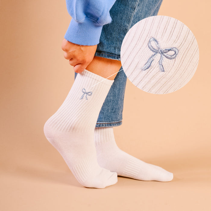 A close-up shows The Darling Effect's Blue Bow Embroidered Crew Socks—white, ribbed, with a blue bow and detailed embroidery, made from breathable fabric. Magnified inset highlights the design. Available in women's sizes.