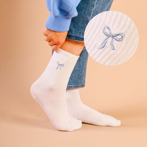 A close-up shows The Darling Effect's Blue Bow Embroidered Crew Socks—white, ribbed, with a blue bow and detailed embroidery, made from breathable fabric. Magnified inset highlights the design. Available in women's sizes.