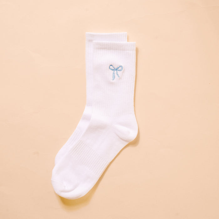 The Darling Effect's Blue Bow Embroidered Crew Socks for women feature delicate embroidery, a light blue bow at the cuff, and breathable fabric; showcased against a soft peach background.