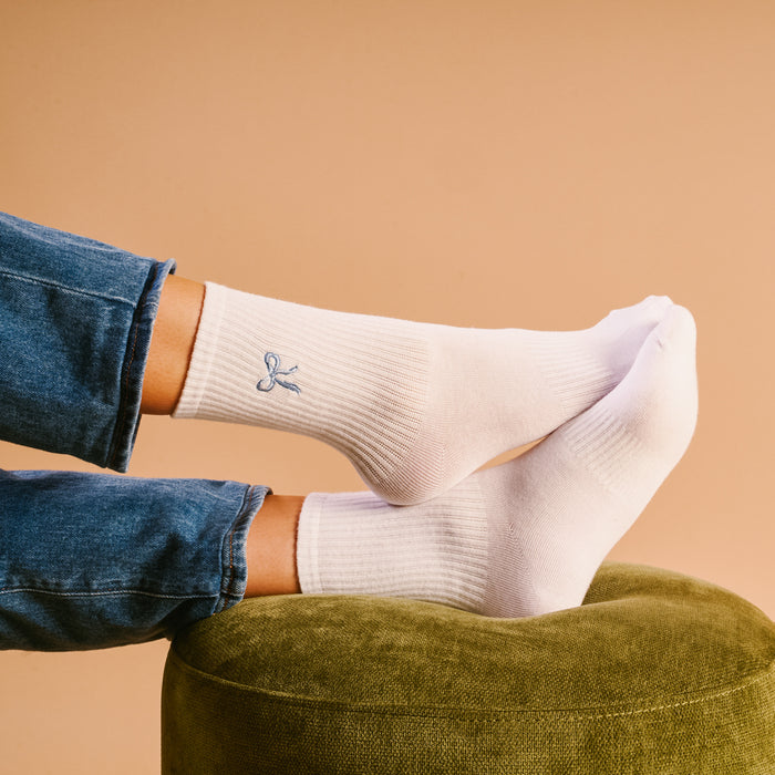 A person wearing The Darling Effect's Blue Bow Embroidered Crew Socks rests their feet on a green ottoman, dressed in blue jeans against a light tan background.