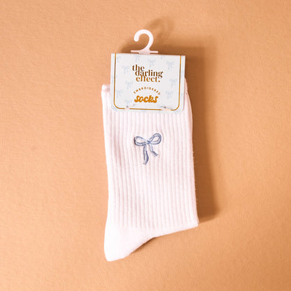 The Darling Effect Blue Bow Embroidered Crew Socks feature white ribbed, breathable fabric with a blue embroidered bow. Tagged and displayed on a white retail hook, these socks are available in women’s sizes.