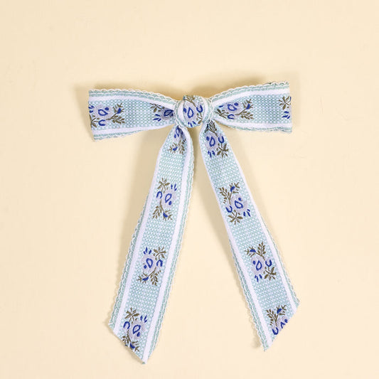 The Darling Effect Blue Heirloom Rosette Hair Bow features a light blue ribbon with floral patterns, tied in a classic bow with two loops and long tails, perfect for hair accessories. Displayed flat on a light beige background.