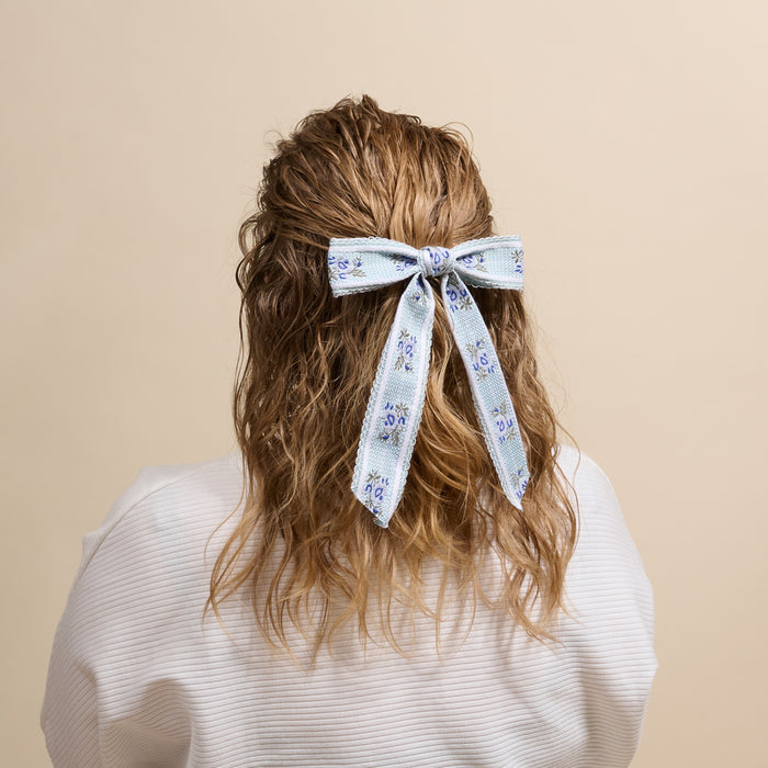 A person with wavy, shoulder-length hair seen from behind, wearing a white top and sporting The Darling Effect's Blue Heirloom Rosette Hair Bow—a striking accessory that adds charm to their hairstyle.