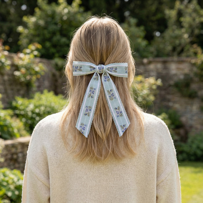 A person with long blonde hair, seen from behind, wears a cream sweater and The Darling Effect's Blue Heirloom Rosette Hair Bow. A garden with greenery and a stone wall appears in the background.