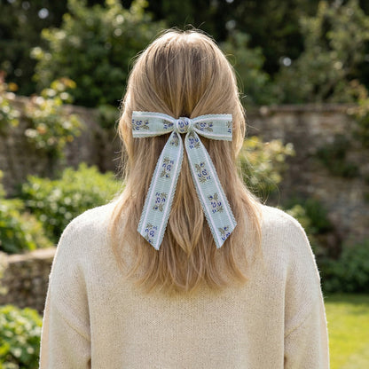 A person with long blonde hair, seen from behind, wears a cream sweater and The Darling Effect's Blue Heirloom Rosette Hair Bow. A garden with greenery and a stone wall appears in the background.