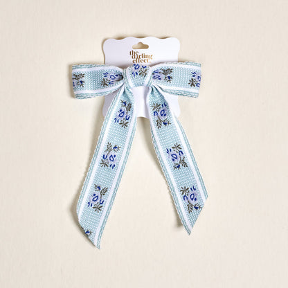The Darling Effect Blue Heirloom Rosette Hair Bow features light blue floral embroidery and comes tied to a scalloped white card with the brand logo, perfect for adding a delicate, textured accent to your favorite hairstyles.