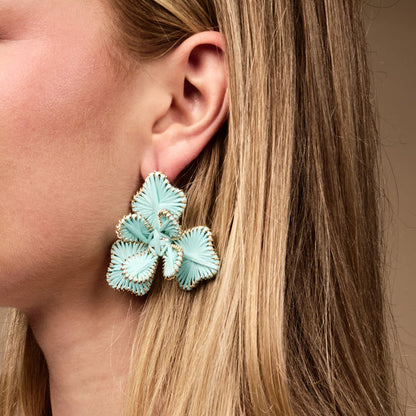 Close-up of a woman wearing The Darling Effect's Blue Main Character Earrings—lightweight, statement boho earrings shaped like light blue flowers with gold accents. Her blond hair is tucked behind her ear, perfect for everyday style.