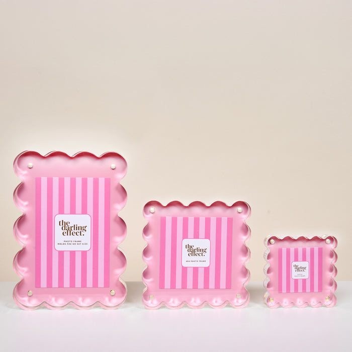 The Blush Little Moments Acrylic Picture Frame by The Darling Effect is shown against a light background, arranged from largest to smallest left to right. It features pink scalloped edges, vertical stripes, and the brand logo in the center.