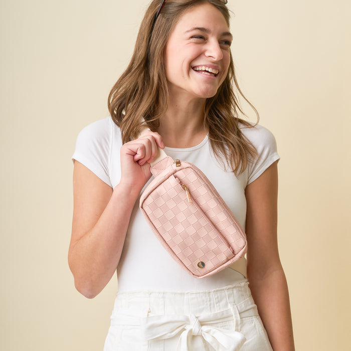 A smiling woman in a white t-shirt and high-waisted white pants holds The Darling Effect's Blush Urban Check Belt Bag, a light pink vegan leather accessory, against a plain beige background.