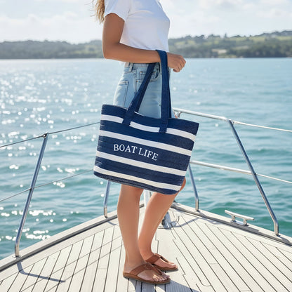 A woman stands on a boat deck by the water, wearing a white tee, denim shorts, sandals, and holding The Darling Effect's Boat Life Straw & Sun Tote. The tote is blue-and-white striped and the sunny shoreline is in the background.