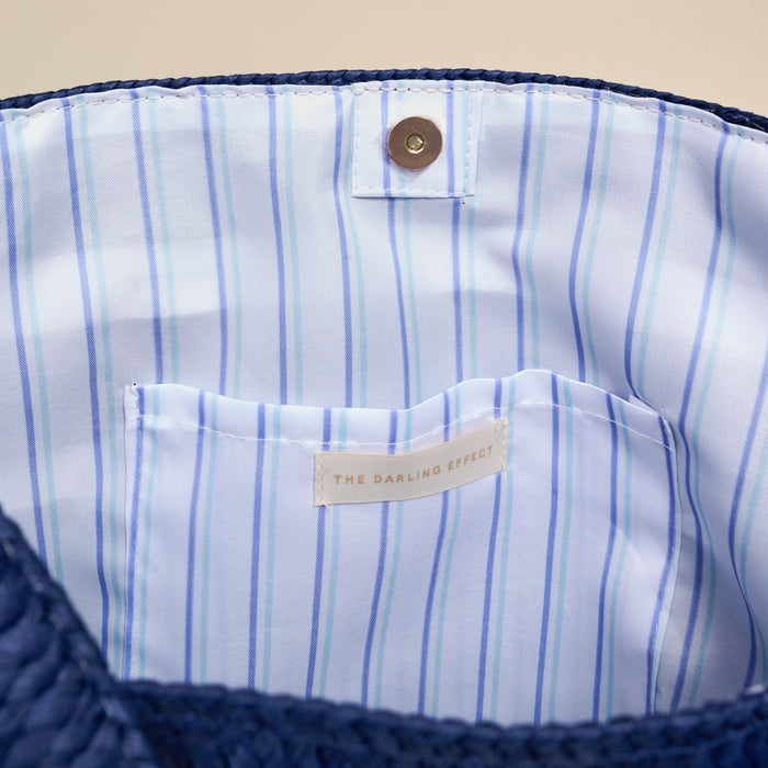 The image shows the inside of The Darling Effect’s Boat Life Straw & Sun Tote, featuring blue woven straw, a light blue and white striped lining, an interior pocket, magnetic snap closure, and a branded label.