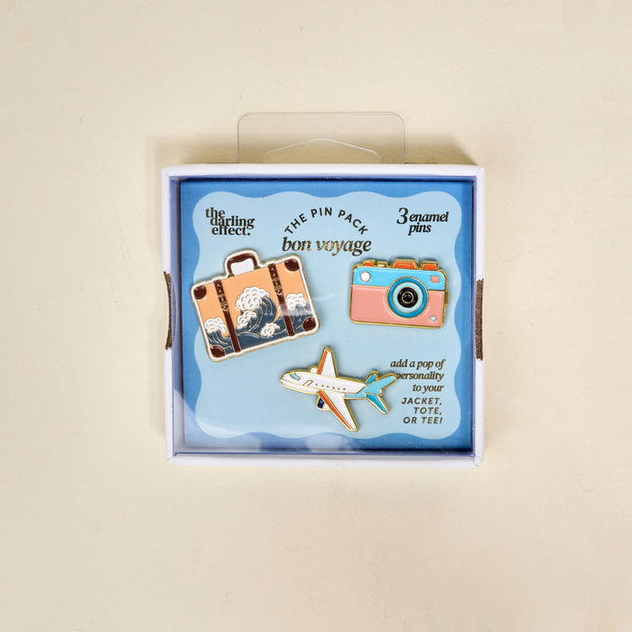 Bon Voyage The Pin Pack by The Darling Effect features three enamel pins—a vintage suitcase, blue camera, and airplane on a blue background—perfect for adding personality to your jacket, tote, or tee!.