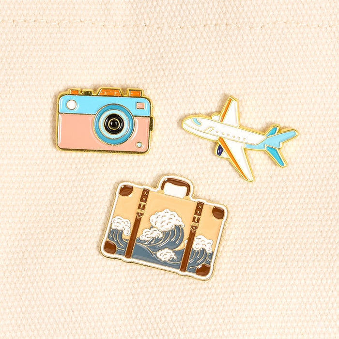 Bon Voyage The Pin Pack by The Darling Effect features three colorful pins—a pink camera, white airplane with blue accents, and a wave suitcase—on textured cream fabric, perfect for any pin lover’s collection.
