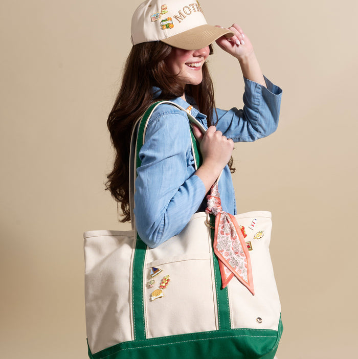 A smiling young woman wears a beige cap with "MOTH" embroidered in gold and collectible pins from The Darling Effect’s Bon Voyage The Pin Pack. She carries a large canvas tote bag adorned with expressive accessories and a floral scarf.