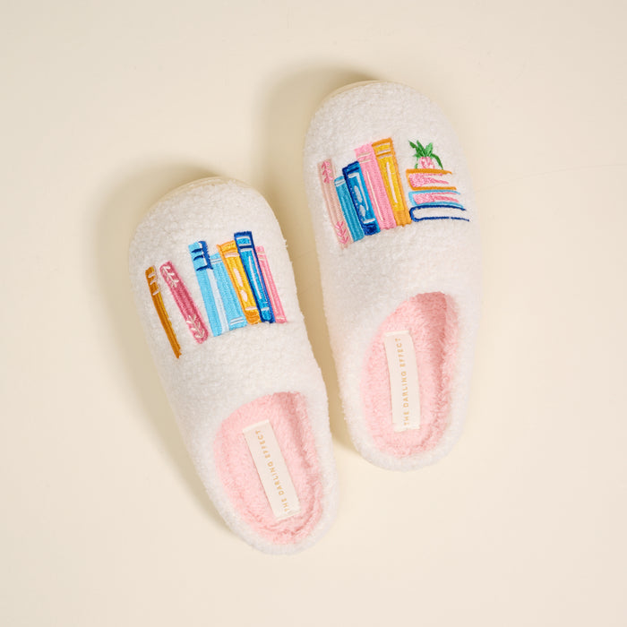 Book Nook Fuzzy Slippers by The Darling Effect: cozy white plush slippers embroidered with vibrant stacks of books and a potted plant.