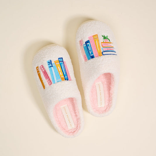 Book Nook Fuzzy Slippers by The Darling Effect: cozy white plush slippers embroidered with vibrant stacks of books and a potted plant.