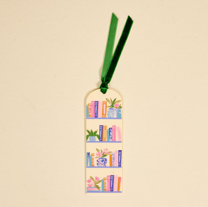 The Book Shelfie Acrylic Bookmark features colorful book and plant illustrations on three shelves against a light background, finished with a green velvet ribbon tassel—perfect for book lovers.
