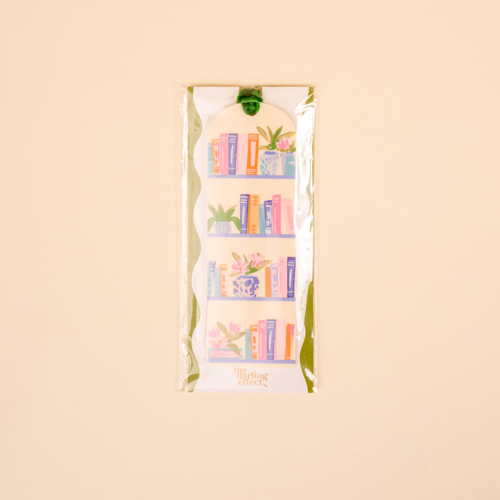 The Darling Effect's Book Shelfie Acrylic Bookmark features a bookshelf of colorful books and plants, packaged in clear wrap with a green velvet ribbon tassel, all on a light beige background—perfect for book lovers.