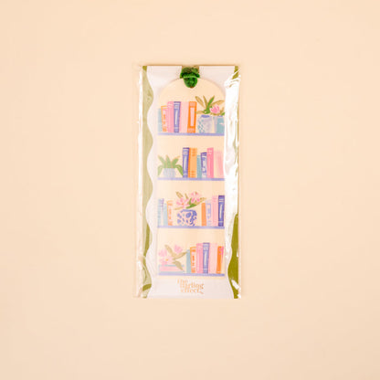 The Darling Effect's Book Shelfie Acrylic Bookmark features a bookshelf of colorful books and plants, packaged in clear wrap with a green velvet ribbon tassel, all on a light beige background—perfect for book lovers.