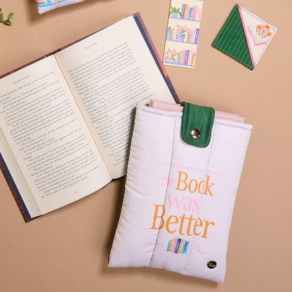 A flat lay photo for book lovers featuring an open book, a padded pink sleeve labeled "The Book Was Better," vibrant Book Shelfie Acrylic Bookmarks with velvet ribbon tassels, and a small plant on a tan surface.
