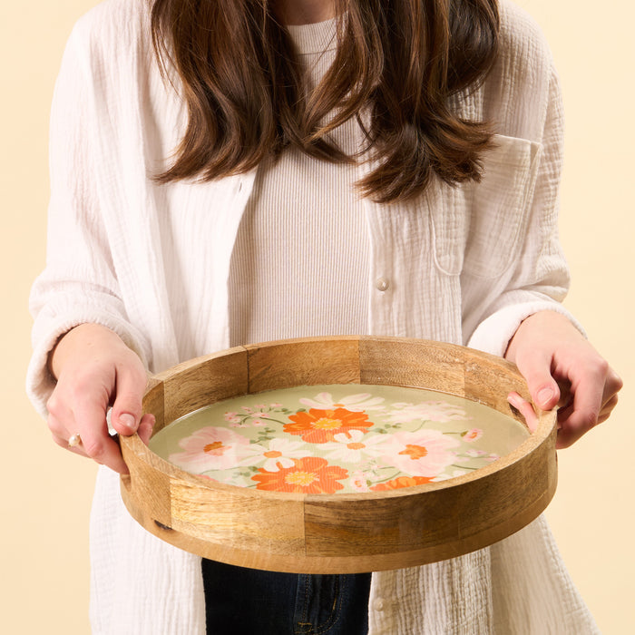 A woman in a white shirt holds the-darling-effect's Bouquet Beauty Sage Large Round Wood Enamel Tray, filled with water and floating orange and white flowers, softly lit against a neutral background.