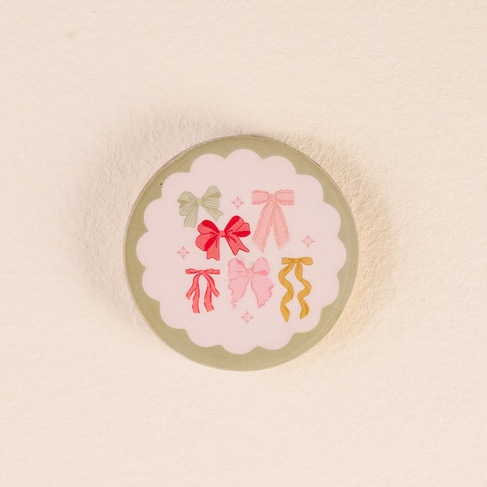 Store your Bow Affair Wood Enamel Coasters from The Darling Effect in style with this round tin, featuring a scalloped white label adorned with six colorful illustrated bows arranged in a circle on a light background.
